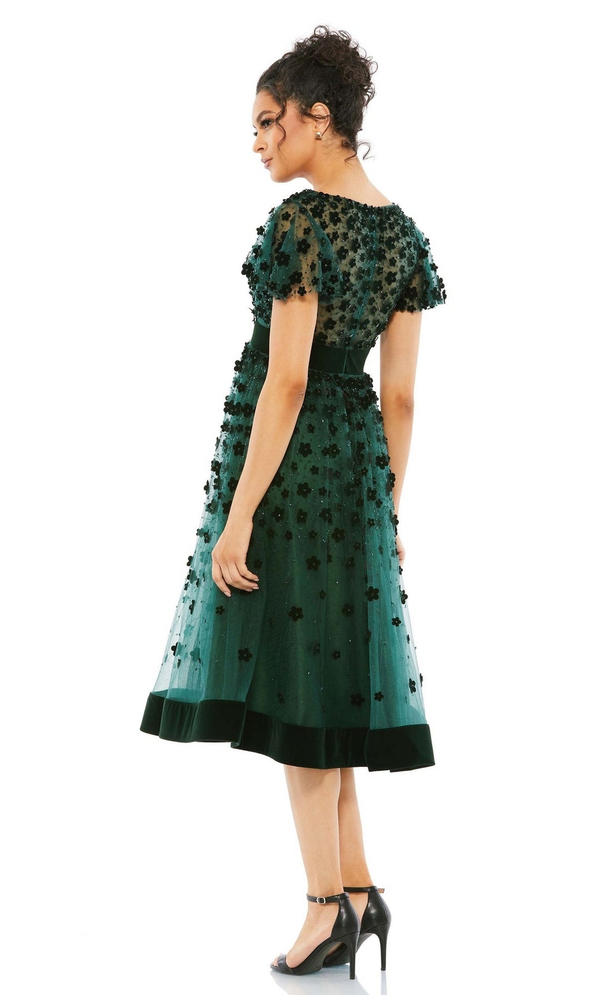 A woman stands in the Mac Duggal 67854 Floral Midi Wedding Guest Dress, featuring green floral embellishments, short sleeves, and a knee-length cut. She completes her vintage look with black ankle-strap heels.